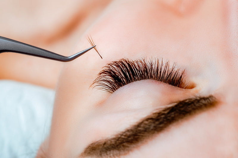 A woman getting hybrid eyelash extensions applied by a lash artist. The lash artist is using tweezers to apply a mix of classic and volume eyelash extensions to the woman's natural lashes