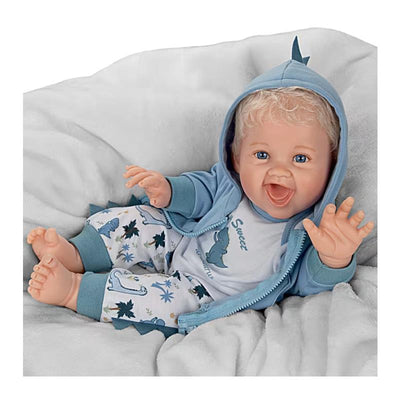 Ashton Drake Dinosaur Baby Boy Doll, wearing a blue hoodie and patterned pants, smiling with arms raised on a soft blanket.