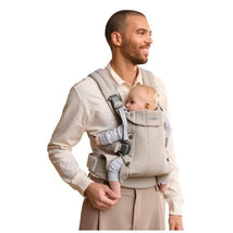 Light beige BabyBjörn Baby Carrier Harmony worn by a smiling man, carrying a baby who is comfortably seated inside.