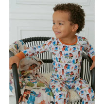 Toddler wearing a colorful Baby Ohana bamboo pajama set featuring various cartoon characters, sitting on a chair with a blanket.