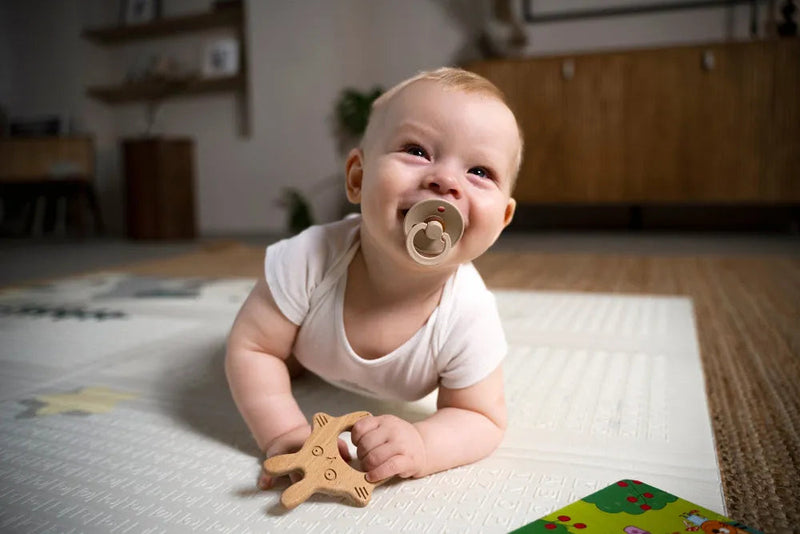 Smiling baby on play mat with wooden teether and pacifier, ideal for infant development and comfort.
