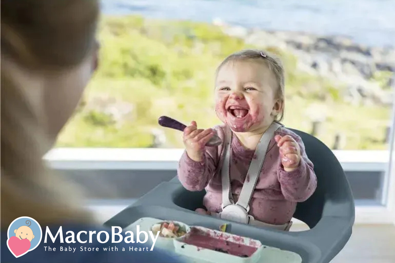 Happy baby in high chair eating messy meal with spoon, smiling at mom
