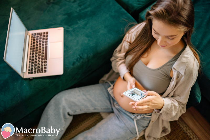 Pregnant woman browsing baby products on phone with laptop nearby, shopping for maternity and baby essentials