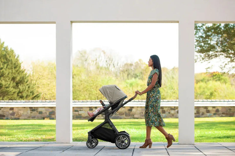 Mom pushing baby stroller outdoors under white archway, green lawn background