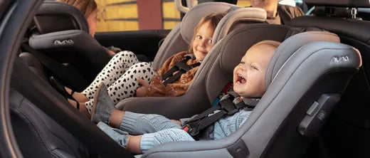 Two happy babies in gray car seats inside a vehicle, smiling and secured with harnesses.
