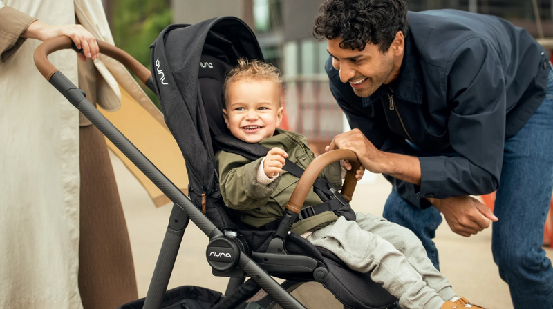Happy baby in black Nuna stroller with parents outdoors, smiling and interacting