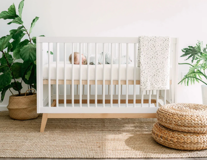 Baby sleeping in white modern crib with speckled blanket, natural rug, and potted plants in nursery