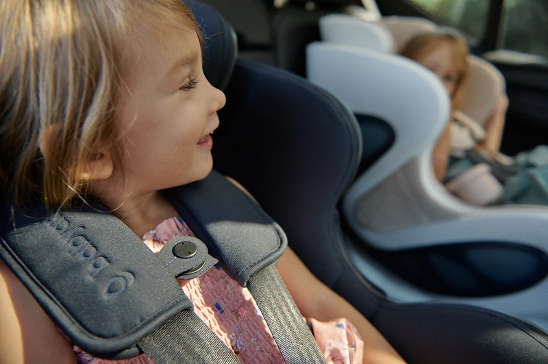 Toddler in black car seat with harness, smiling beside another child in light gray car seat