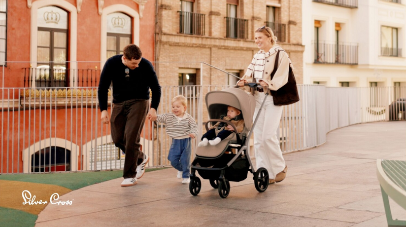 Family walking outdoors with baby in stroller, toddler holding dad's hand, urban setting