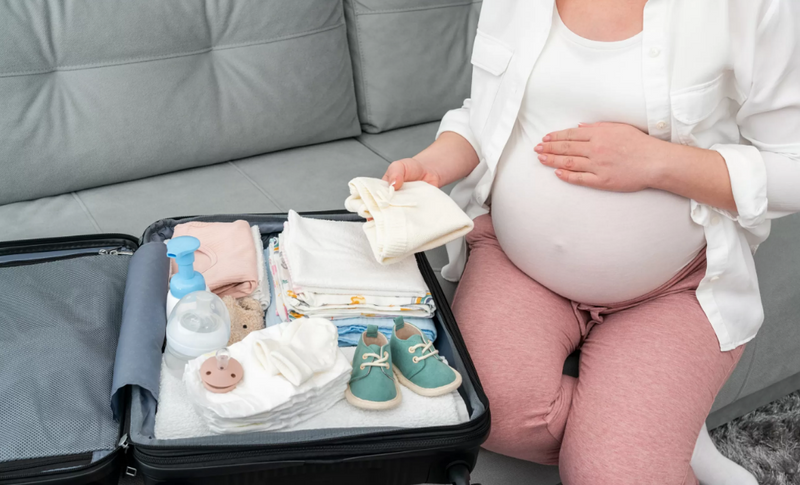 Pregnant woman packing baby essentials like clothes, shoes, and diapers into suitcase for hospital stay