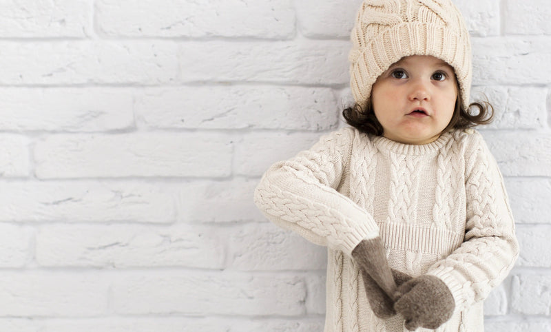 Toddler in cozy cream cable knit sweater, hat, and mittens against white brick wall