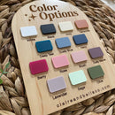 Wooden color swatch board with 14 labeled paint samples for baby products, including White, Black, Navy, Rose, Sage, Olive