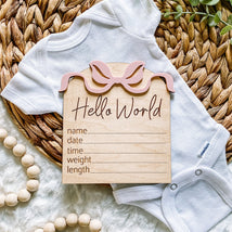 Hello World baby announcement card with pink bow, white onesie, and wooden beads on woven mat