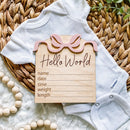 Hello World baby announcement card with pink bow, white onesie, and wooden beads on woven mat