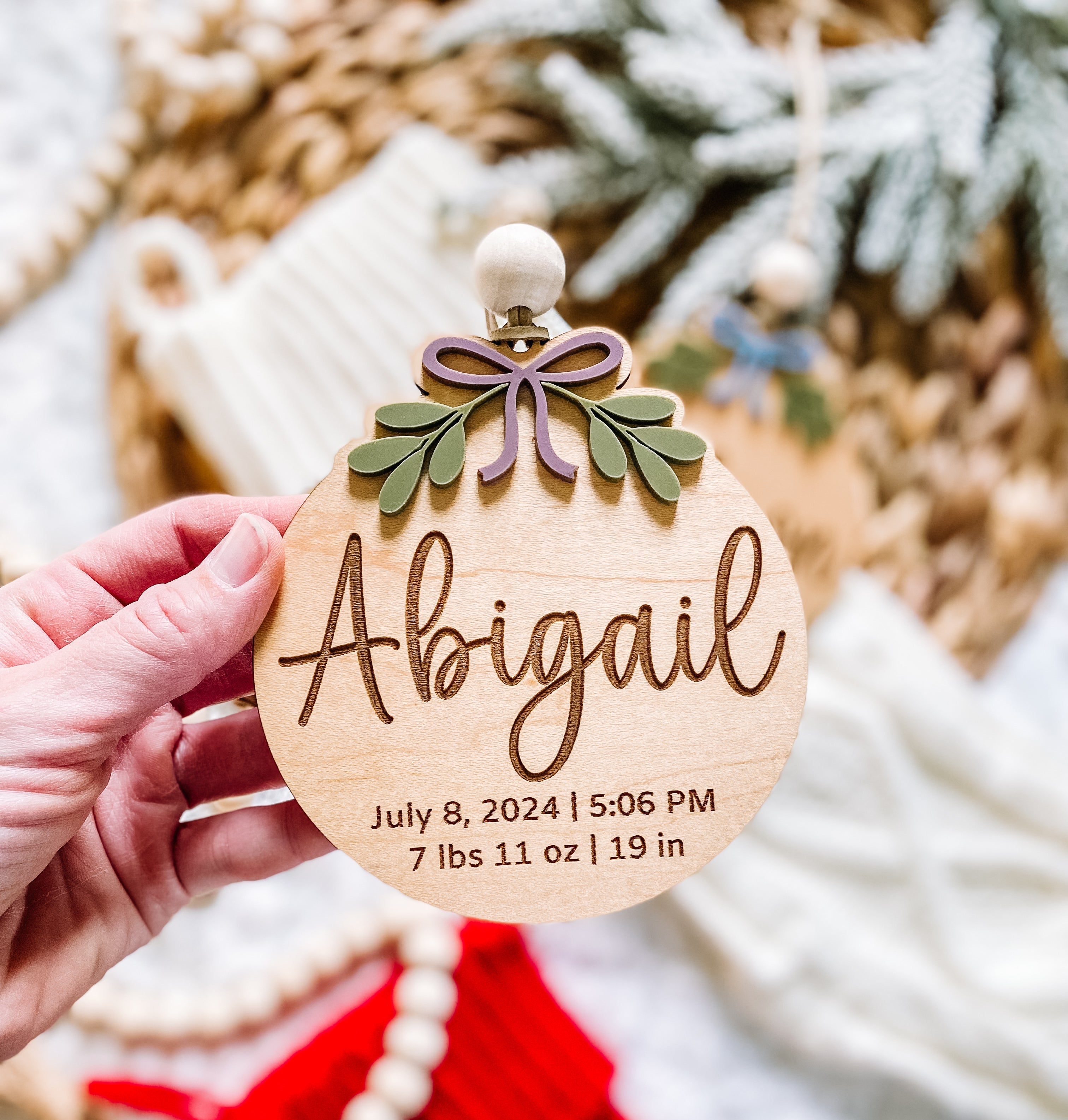 Personalized wooden baby ornament with name Abigail, birth stats, purple bow, and green leaves, held near festive decor