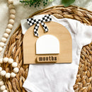 Wooden baby monthly milestone sign with checkered bow, on white onesie, wicker basket, baby clothing