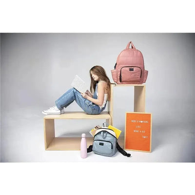 Diaper backpack in rose dawn color displayed next to a model reading, with a gray backpack and water bottle nearby.