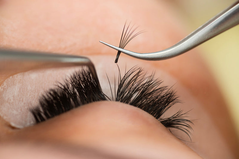 A close-up of a woman getting volume eyelash extensions applied by a lash artist. The lash artist is using tweezers to apply eyelash fans extensions to each of the woman's natural lashes
