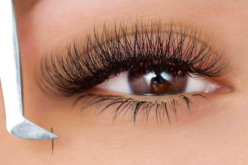 A close-up of a woman getting Brazilian volume eyelash extensions applied by a lash artist. The lash artist is using tweezers to apply multiple ultra-fine eyelash fans extension to each of the woman's natural lashes