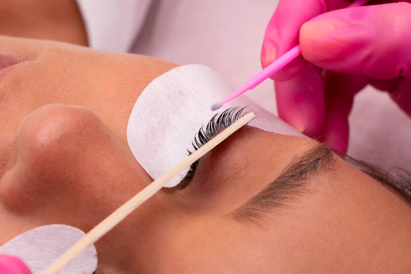 A close-up of a woman getting her eyelash extensions removed by a lash artist. The lash artist is using a cotton swab to apply a remover to the woman's eyelashes