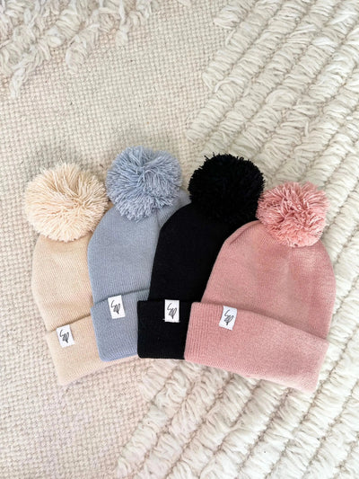 Four Sweet P Baby Co. pom pom beanies in cream, light blue, black, and pink on a white rug.