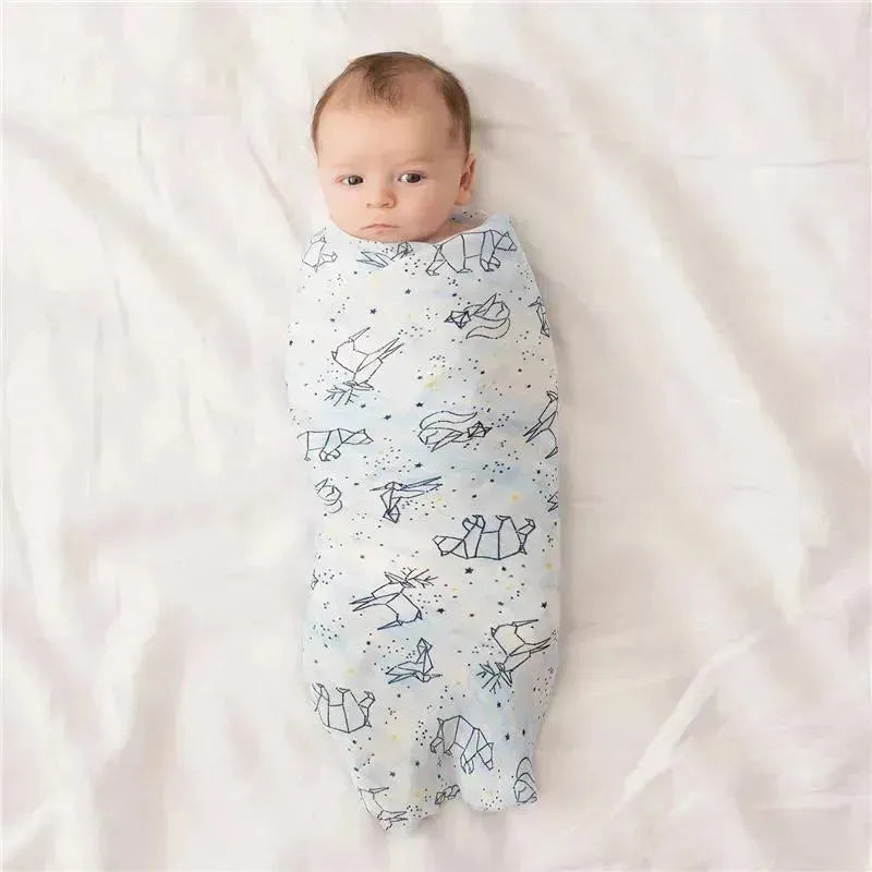 A wrapped baby lying on a white surface, swaddled in a light blue Aden + Anais fabric featuring stargaze designs.