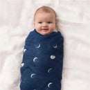 Baby wrapped in a silky soft navy aden + anais swaddle with moon patterns, lying on a light blanket and smiling.