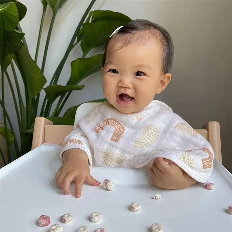 Smiling baby wearing an Aden + Anais Keep Rising Burpy Bib, sitting at a table with snacks scattered on the surface.