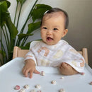Smiling baby wearing an Aden + Anais Keep Rising Burpy Bib, sitting at a table with snacks scattered on the surface.