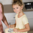 Young girl in a yellow dress smiles while holding a gray bowl and spoon, engaged in feeding a doll with colorful accessories.