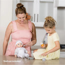 A woman and a girl play with a doll accessory set; the woman feeds a toy doll while the girl holds a feeding bottle.