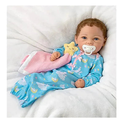 Baby doll Rylee in blue pajamas with cloud and star design, holding a pink blanket and pacifier, lies on a soft surface.