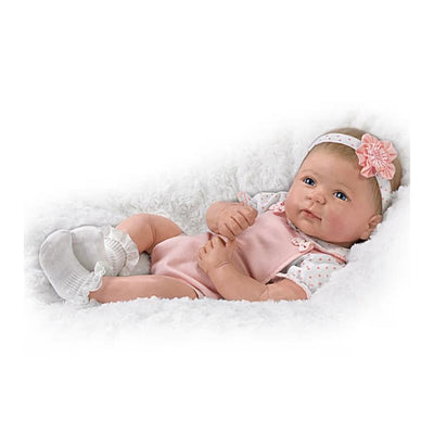 Lifelike silicone baby doll named Ava by Ashton Drake, wearing a pink outfit with a floral headband, resting on soft white bedding.