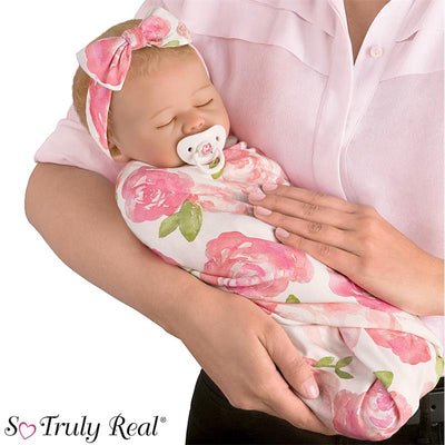 Rosie Baby Girl Doll by Ashton Drake wrapped in a floral swaddle, held by a caregiver with a pacifier in its mouth.