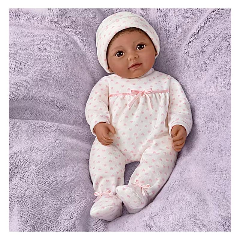 Lifelike Ashton Drake Tiny Miracles 10 Little Kiara doll, dressed in a pink heart-patterned onesie and matching hat, on a soft purple blanket.