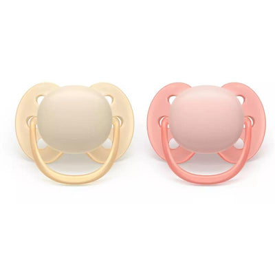 Avent 2Pk Ultra Soft Pacifier set for 0-6 months, featuring a cream and a soft pink pacifier with rounded nipples and safety handles.
