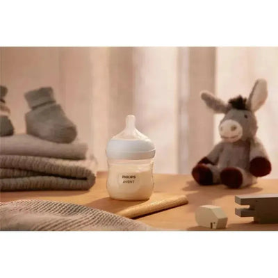 Clear 4oz Philips Avent baby bottle with natural response nipple, placed on a wooden surface with socks and stuffed toys in the background.