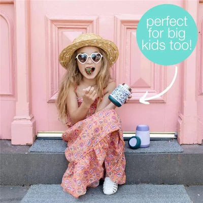 Girl in a pink dress and straw hat eating from a B.box Friendly Fox insulated food jar, with a playful expression and colorful door background.