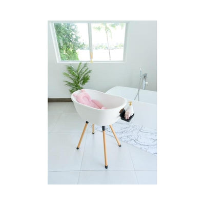 Bath support pillow in petal pink, positioned in a bright bathroom with wooden legs and a nearby plant.