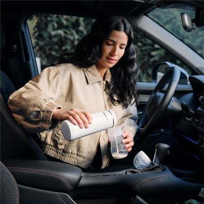 A woman with long dark hair pours milk from a Baby Brezza portable bottle warmer into a baby bottle while sitting in a car.