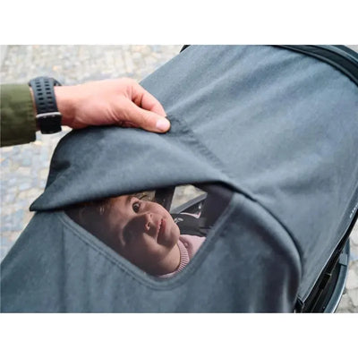 A hand lifts the cover of a Baby Jogger City Prix™ Jogger X, revealing a small child inside looking upwards.