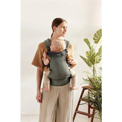 BabyBjorn Baby Carrier Harmony in dark green, featuring a child comfortably seated, worn by a woman with a light brown sweater and striped pants.