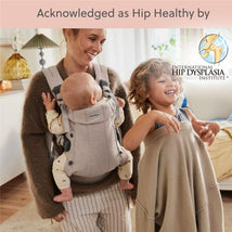 A woman smiles while carrying a baby in a light beige BabyBjörn Baby Carrier, as a young child plays beside them.