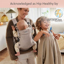A woman smiles while carrying a baby in a light beige BabyBjörn Baby Carrier, as a young child plays beside them.