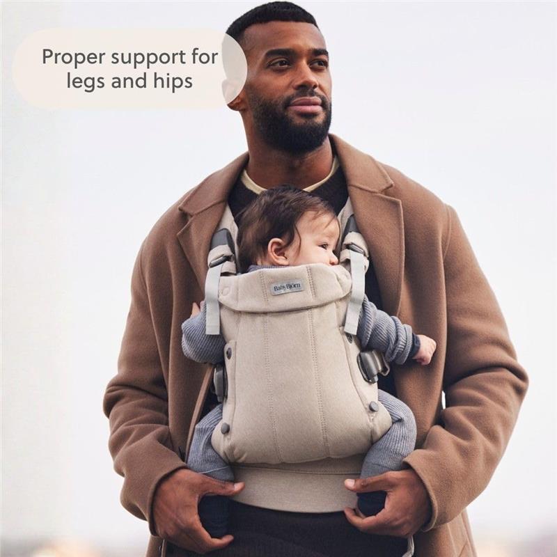 A man wearing a BabyBjörn carrier in light beige with a baby, demonstrating proper support for legs and hips.