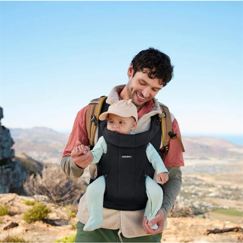 A smiling man wears a Babybjorn Baby Carrier Hold Mesh in black, carrying a baby dressed in light blue outdoors with a scenic background.