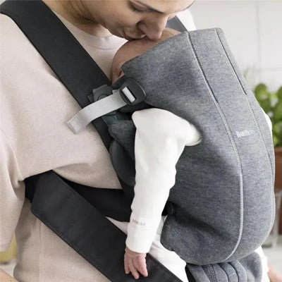 A parent kisses their baby, who is securely nestled in a BabyBjorn Baby Carrier Mini 3D Jersey in dark gray.