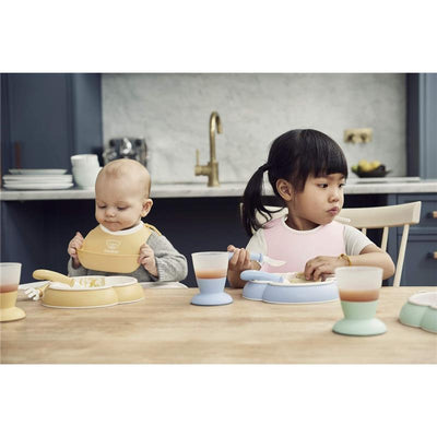 Two children sitting at a wooden table, using Babybjorn products, with the girl eating from a blue plate and the boy in a yellow bib.