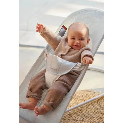 Smiling baby in a BabyBjorn Bouncer Balance Soft Mesh, wearing a beige sweater and brown pants, with arms raised joyfully.