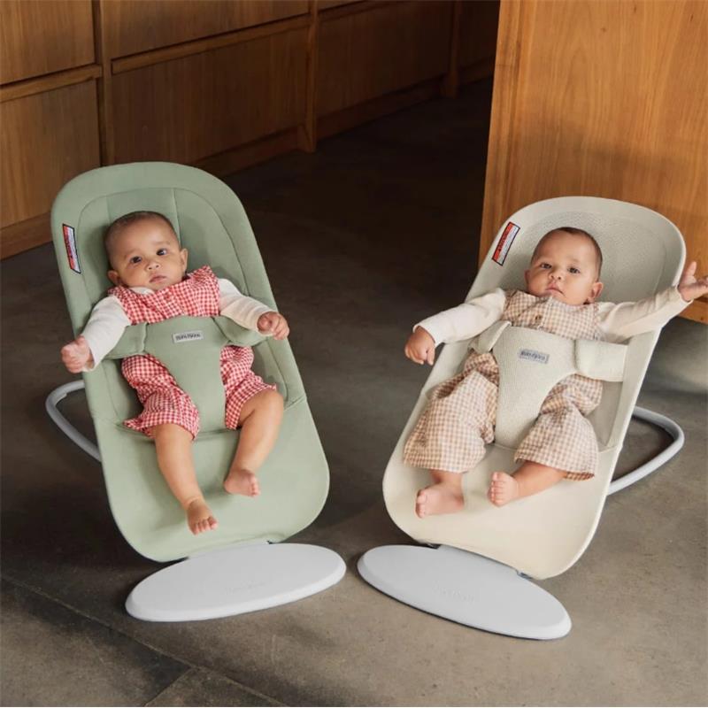 Two infants sitting in Babybjorn Bouncer Begin seats; one has a light green fabric, the other a neutral tone, both feature adjustable harnesses.
